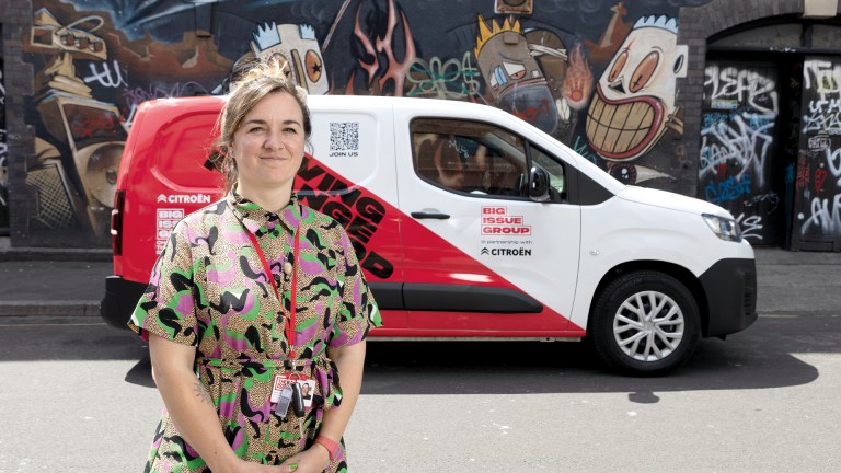 Big Issue frontline manager Hattie Greenyer standing with a Big Issue magazine in front of the Citroën ë-Berlingo Van displaying 'Driving Change for Good' branding, with colourful street art mural in background