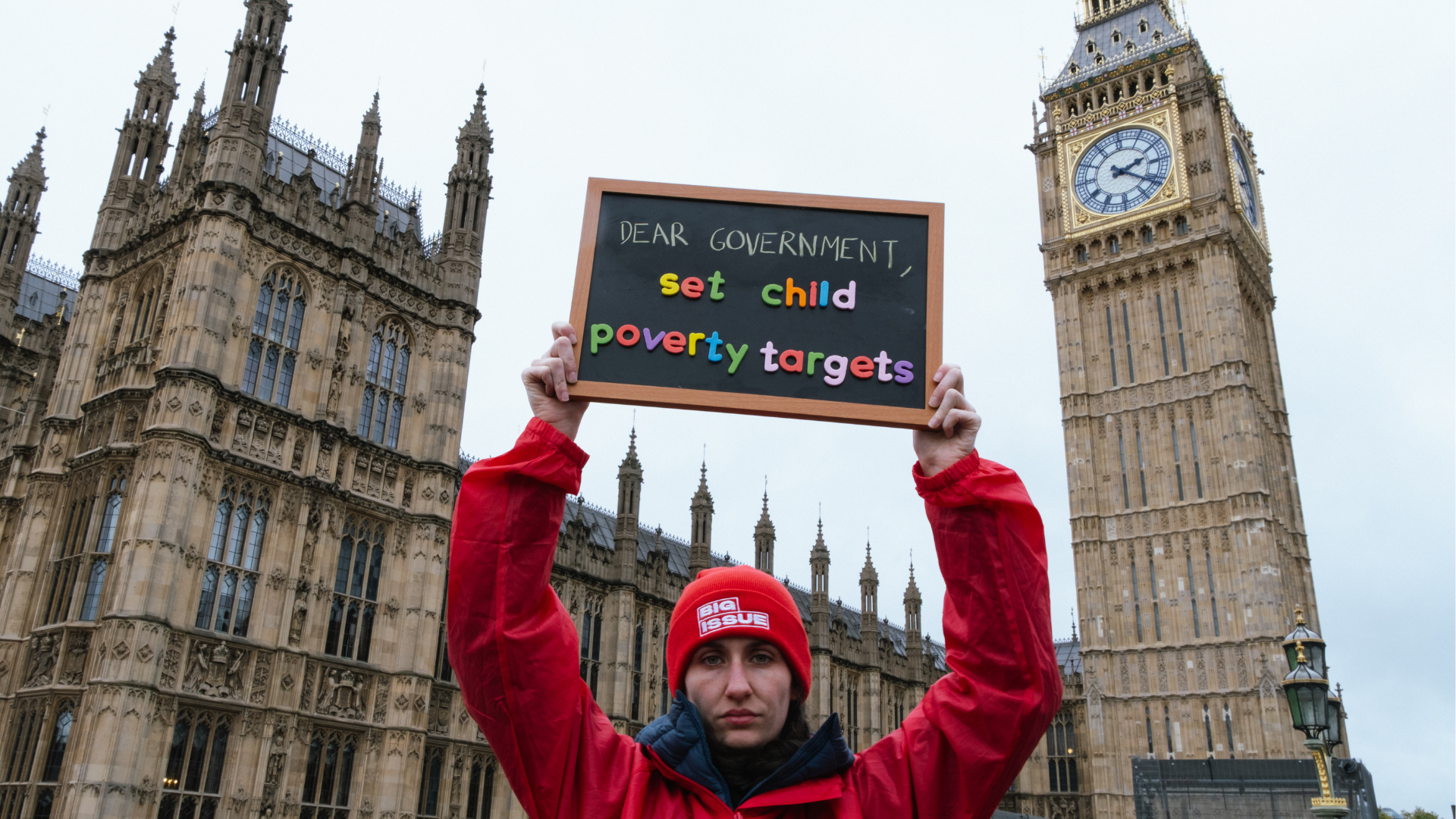 A woman in a Big Issue coat and beanie holds up a sign reading "Dear government: We need child poverty targets" outside the Palace of Westminster in London.