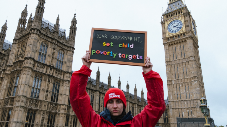 A woman in a Big Issue coat and beanie holds up a sign reading