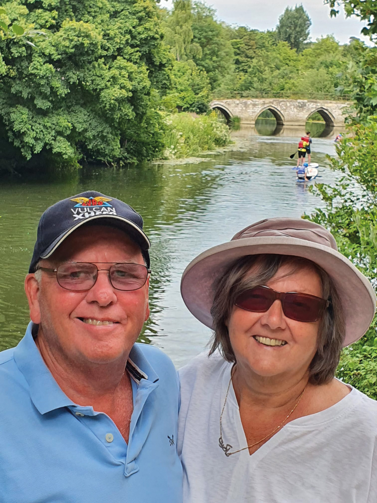 A smiling older couple wearing sunglasses and sun hats pose together beside a calm river, with trees, paddleboarders and a stone arched bridge in the background.