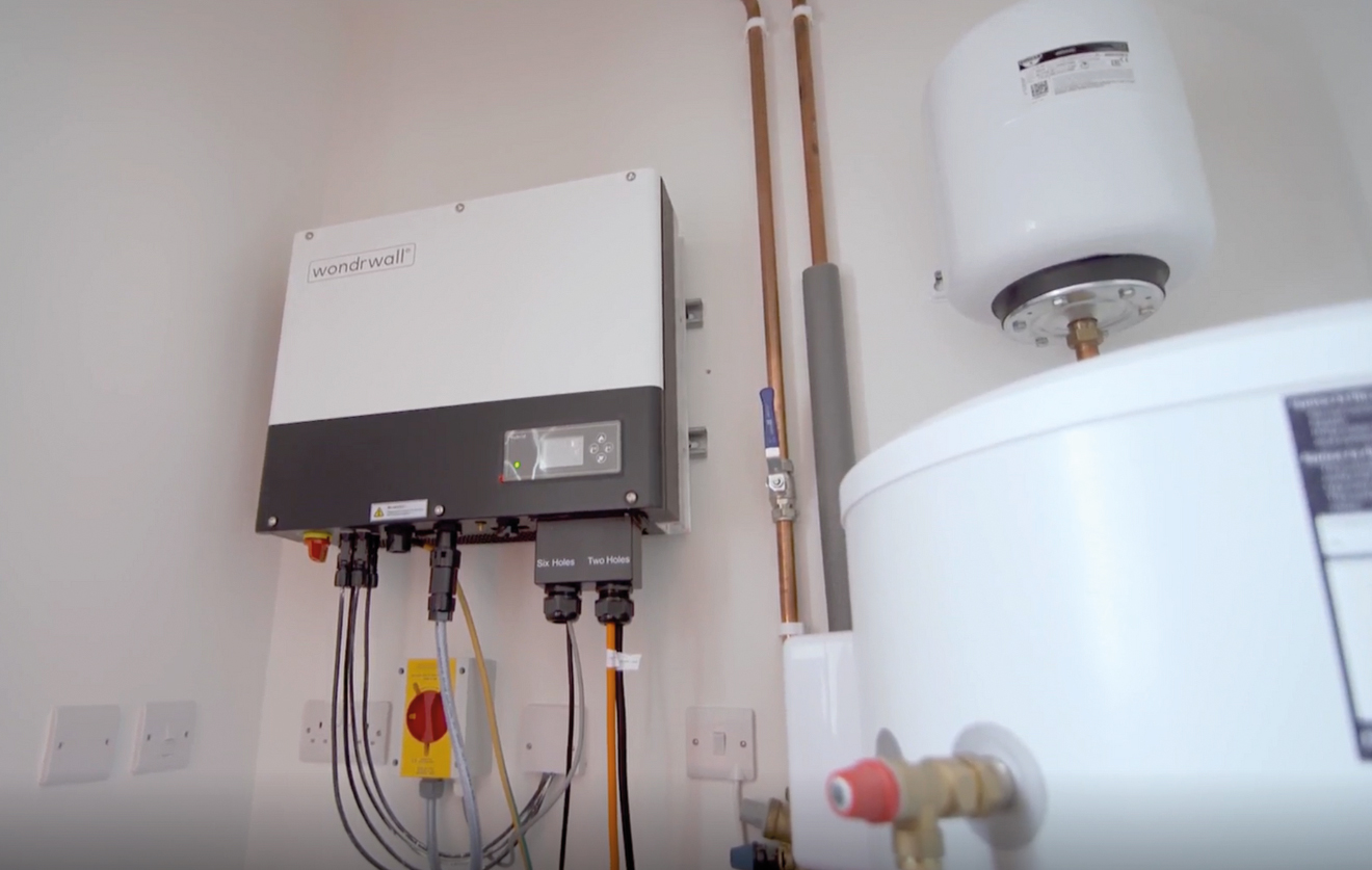 A Wondrwall smart home control unit mounted on a wall alongside copper pipes, electrical cables and a white hot water cylinder in a utility room.
