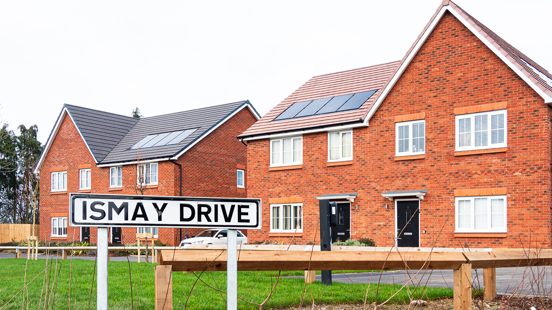 A row of modern red-brick semi-detached houses fitted with rooftop solar panels, photographed behind a street sign reading “Ismay Drive”.