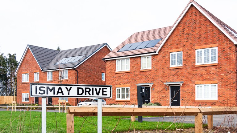 A row of modern red-brick semi-detached houses fitted with rooftop solar panels, photographed behind a street sign reading “Ismay Drive”.