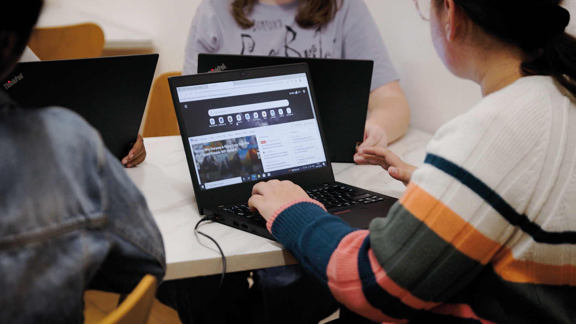 A group of young people sitting around a table using Lenovo ThinkPad laptops, with one screen open on a web browser during a session or workshop.