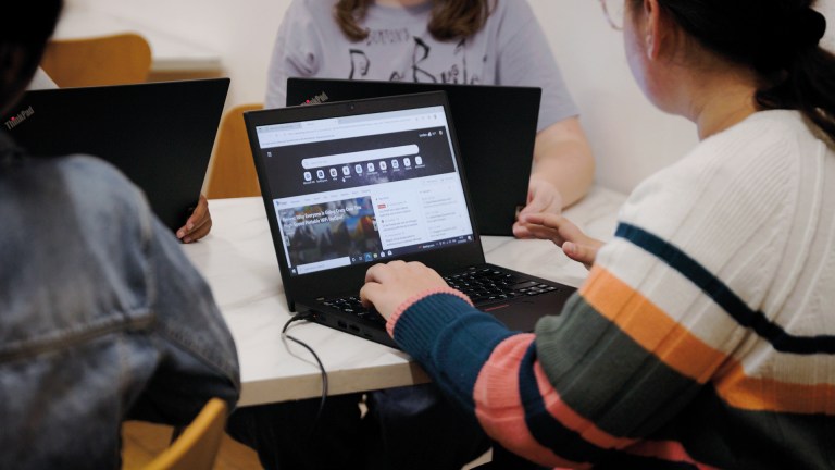 A group of young people sitting around a table using Lenovo ThinkPad laptops, with one screen open on a web browser during a session or workshop.