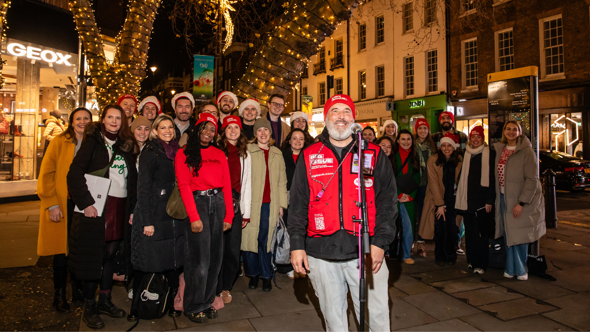 Big Issue vendor Andre Rostant and the Adam Street Singers smile at the camera at their performance spot in Covent Garden, decorated with Christmas lights