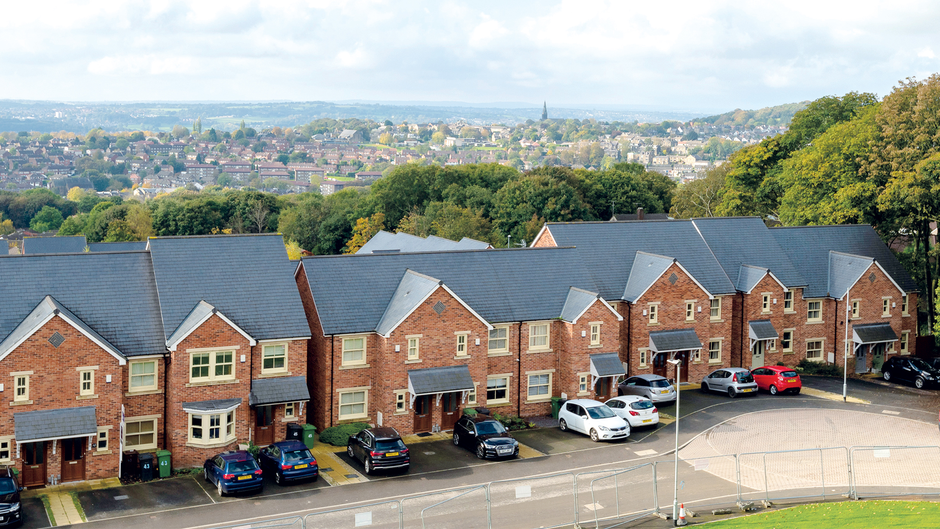 A row of newly built brick homes in a social housing development, with parked cars in front and a town and green hillside visible in the background.