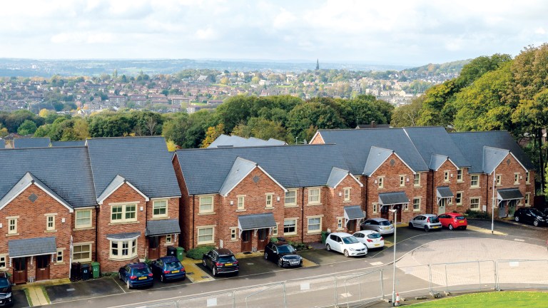A row of newly built brick homes in a social housing development, with parked cars in front and a town and green hillside visible in the background.