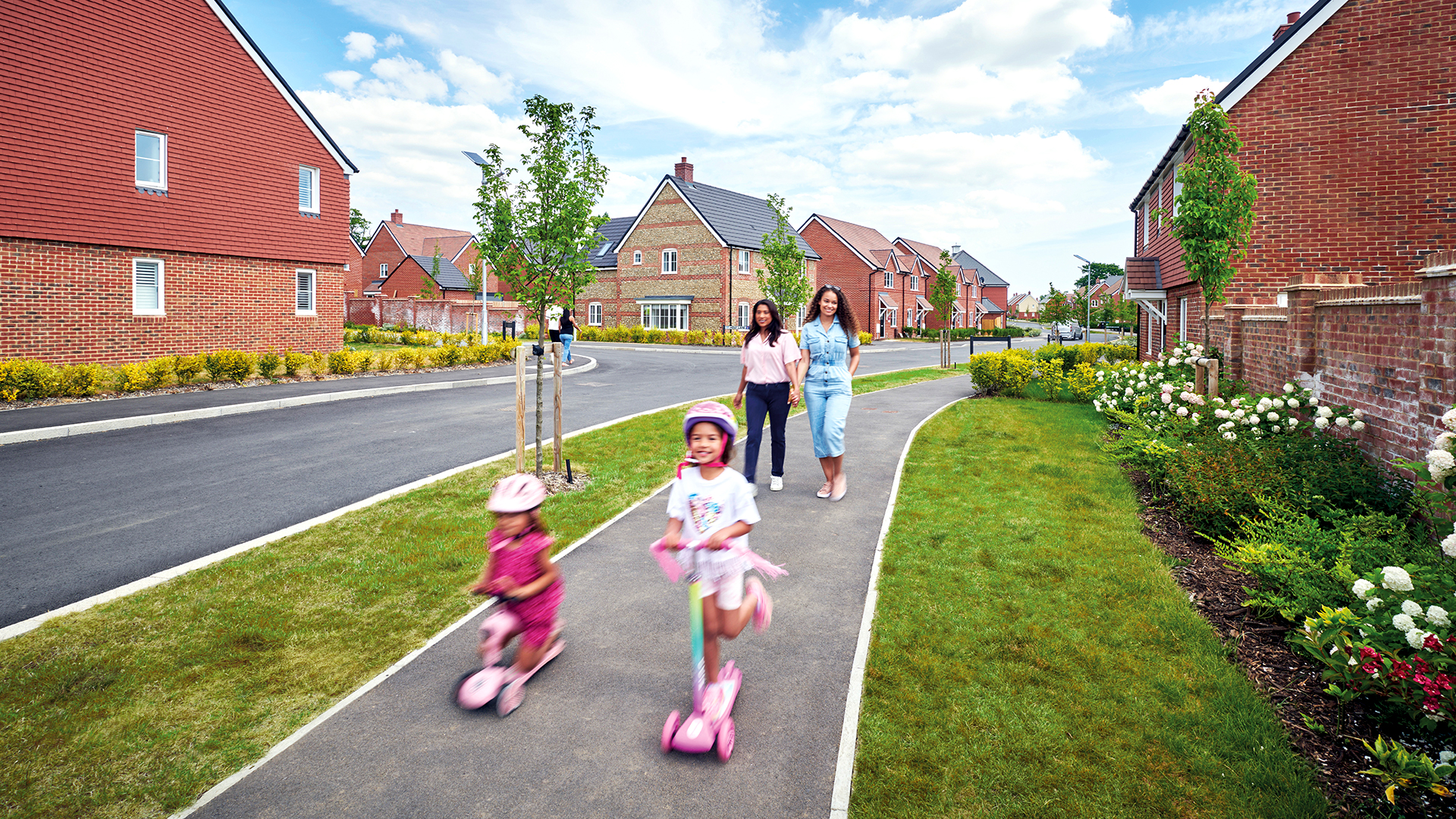Two children riding pink scooters along a paved path in a newly built housing development, with two adults walking behind them and modern brick homes and green spaces on either side.