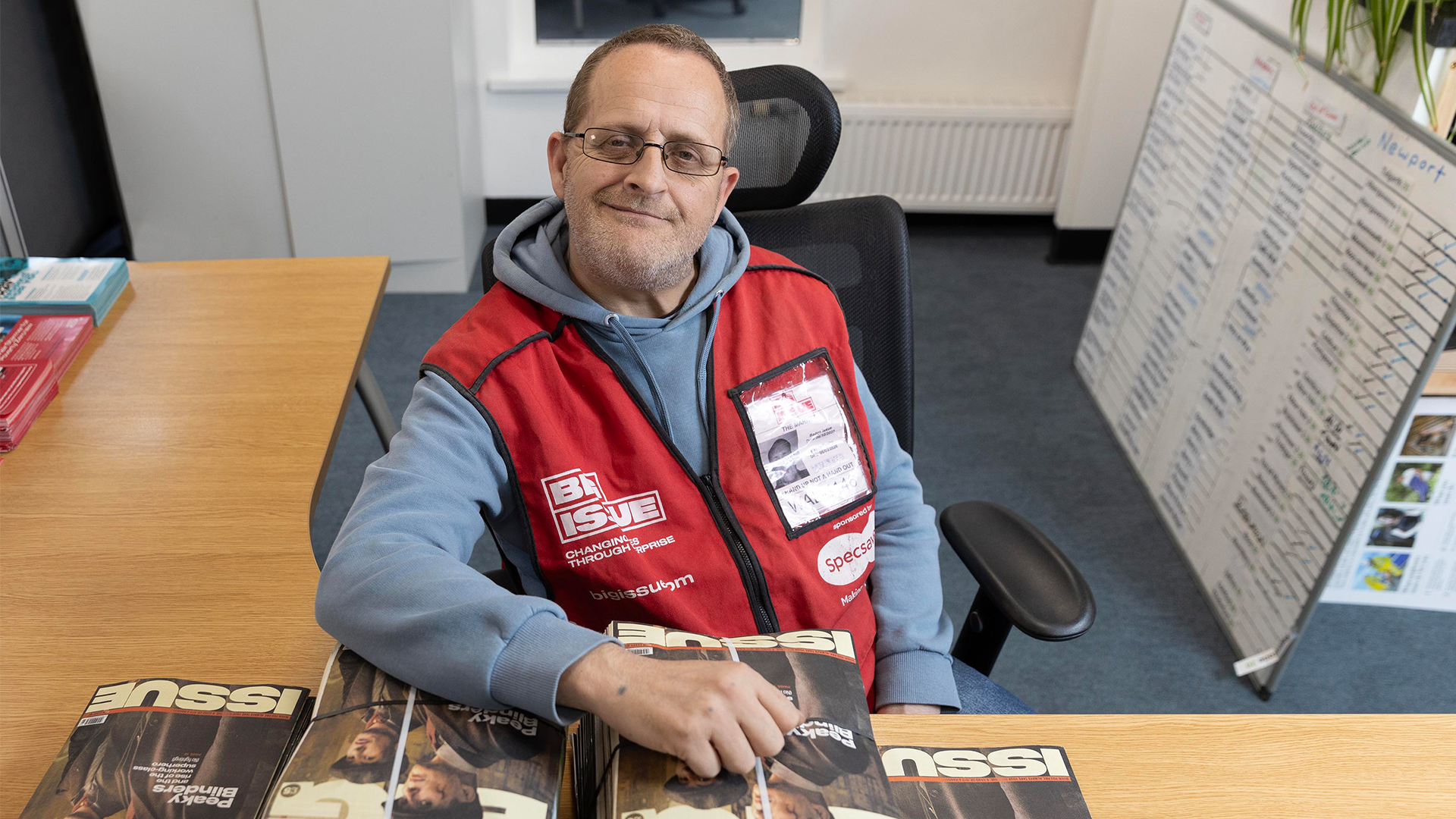 Vendor Stuart sat at a desk, smiling at the camera, wearing a red Big Issue tabard, his arm extended over copies of the magazine
