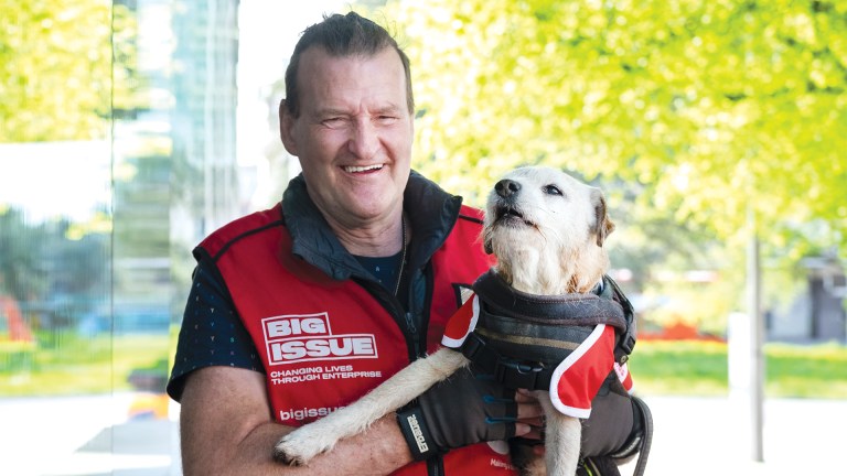 Portrait of Big Issue vendor Clive Rowe holding his dog Geezer. Both are facing the camera outdoors in bright sunlight, with Geezer looking upwards.