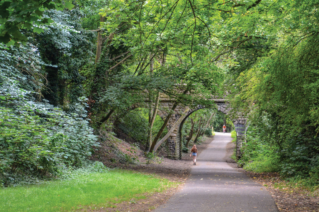 A leafy traffic-free path passes beneath a stone railway bridge on the Two Tunnels Greenway near Bath, with walkers and cyclists in the distance.