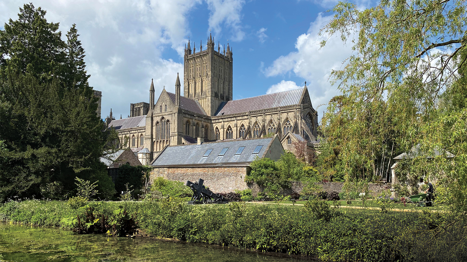 Wells Cathedral seen across the gardens and moat of the Bishop’s Palace beneath a blue, partly cloudy sky.
