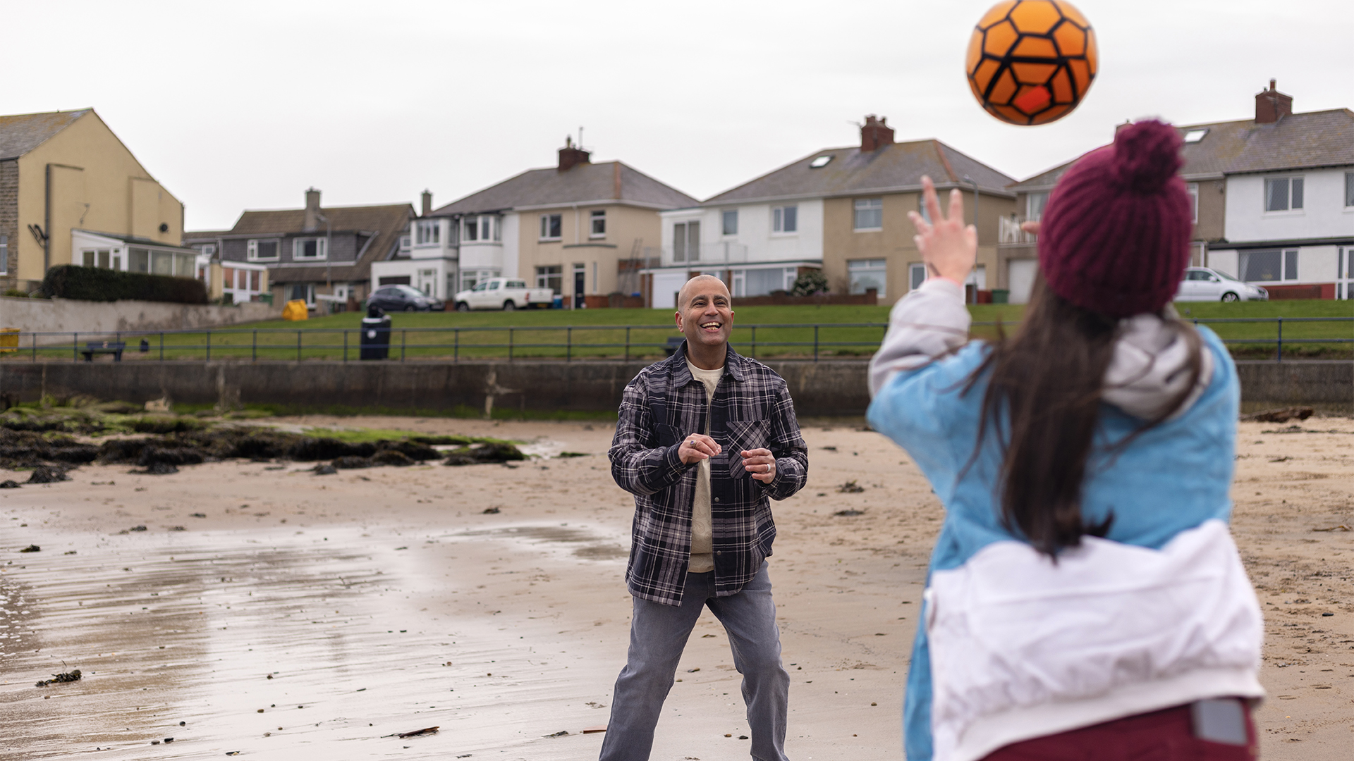 A man and a child play with an orange football on a beach, with houses in the background.